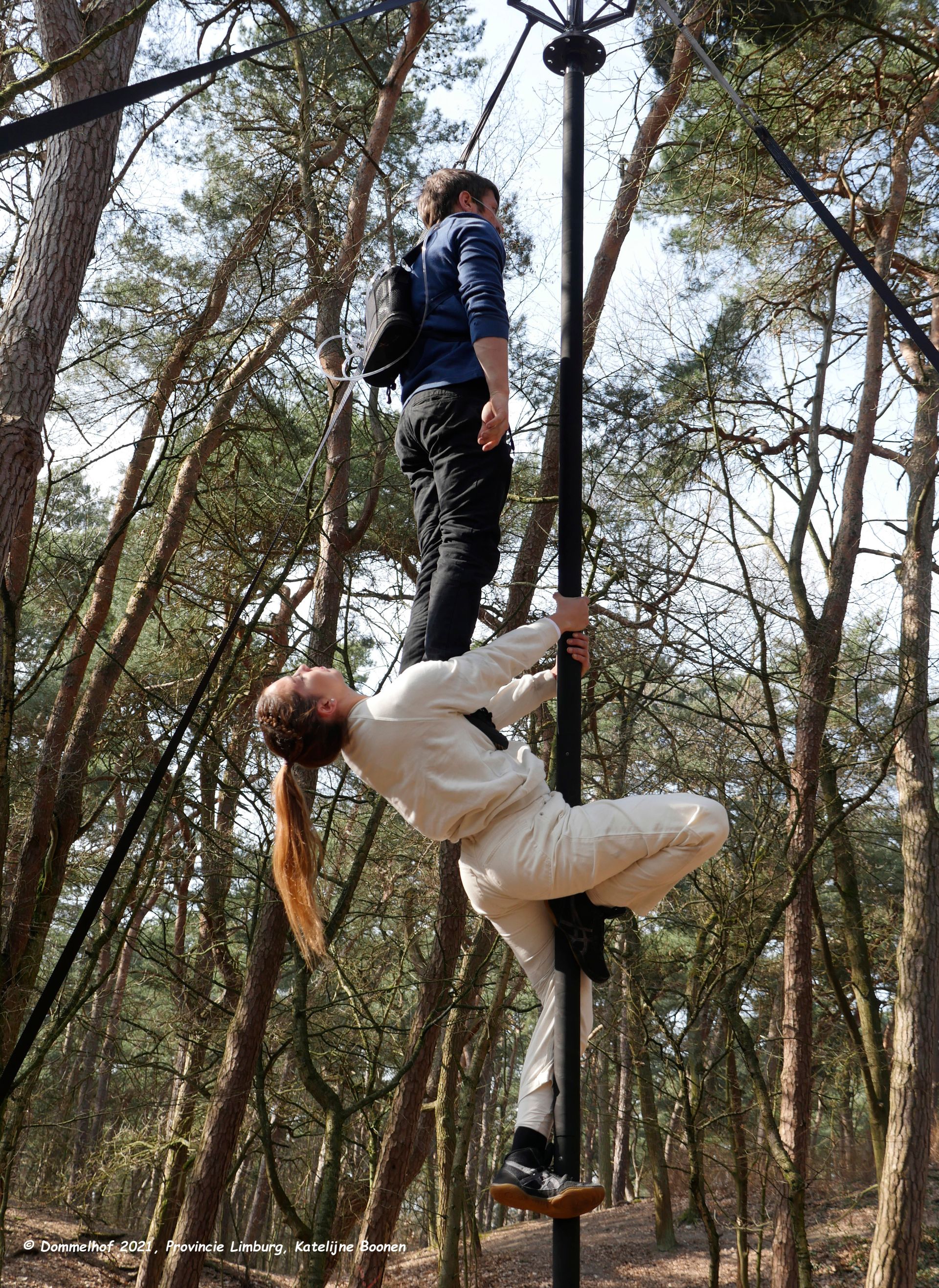 Foto von zwei Artist*innen am Chinesischen Mast. Die untere Person hat Beine und Arme am Mast fest verankert, damit die obere Person auf ihrem Brustkorb aufrecht stehen kann. Die obere Person trägt eine Sauerstofflasche auf dem Rücken.