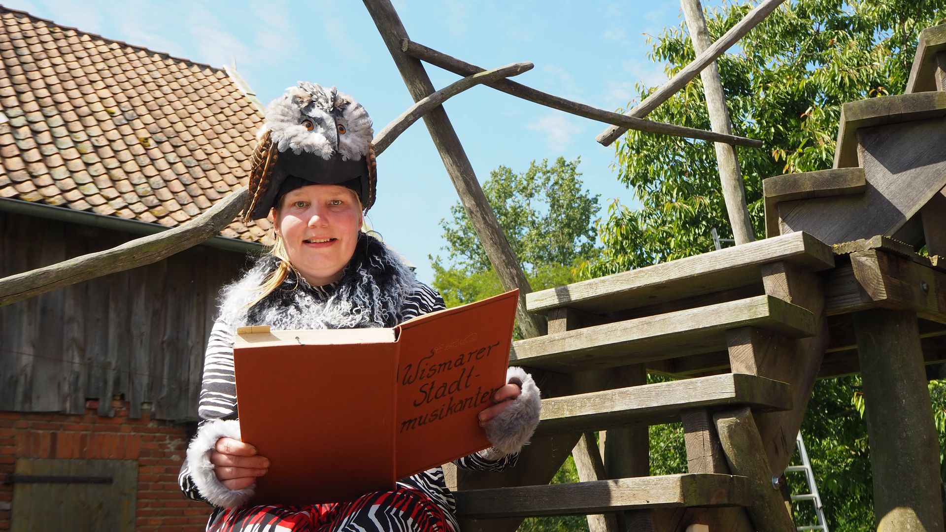 Foto einer Performerin, die auf einer Holztreppe sitzt. Sie hält ein aufgeschlagenes Buch, auf dem steht: "Wismarer Stadtmusikanten". Der Hut auf ihrem Kopf hat die Form einer Eule.
