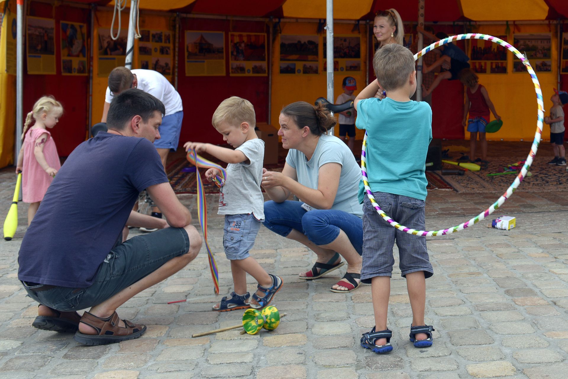 Fotoauszug, auf dem sich ein Kind mit Hoola-Hoop Reifen, eines mit Luftschlangen und einem Diablo sowie ein Kind mit einem Jonglage-Kegel befinden. Im Hintergrund ist ein Pavilion in Form eines Zirkuszelts zu sehen.