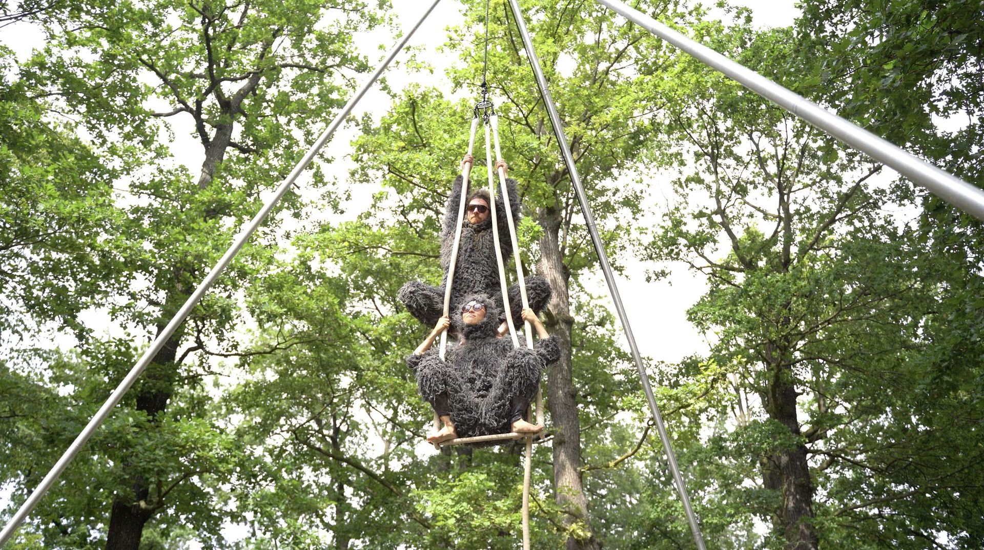 An einer dreibeinigen Traverse hängt ein Trapez. Auf dem Trapez hockt ein*e als Faultier verkleidete Artist*in. Auf den Schultern dieser Artist*in sitzt eine zweite Person in gleichem Kostüm, ein Ganzkörperanzug aus grauem künstlichem Fell.