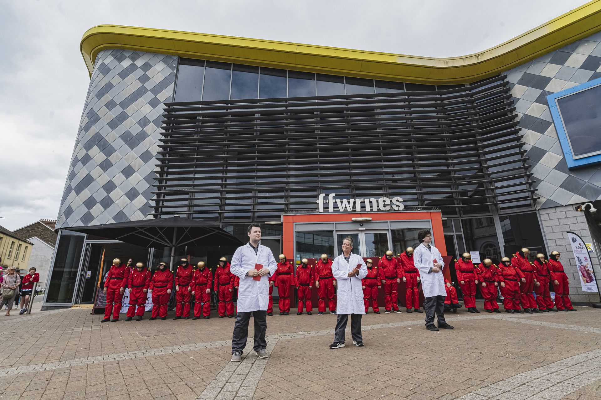 Foto von einer langen Reihe Roboter in roten Overalls vor einem öffentlichen Gebäude. Da die Roboter die Köpfe hängen lassen, scheinen sie ausgeschaltet zu sein. Im Vordergrund stehen drei Personen mit weißen Kitteln, wahrscheinlich Wissenschaftler*innen.