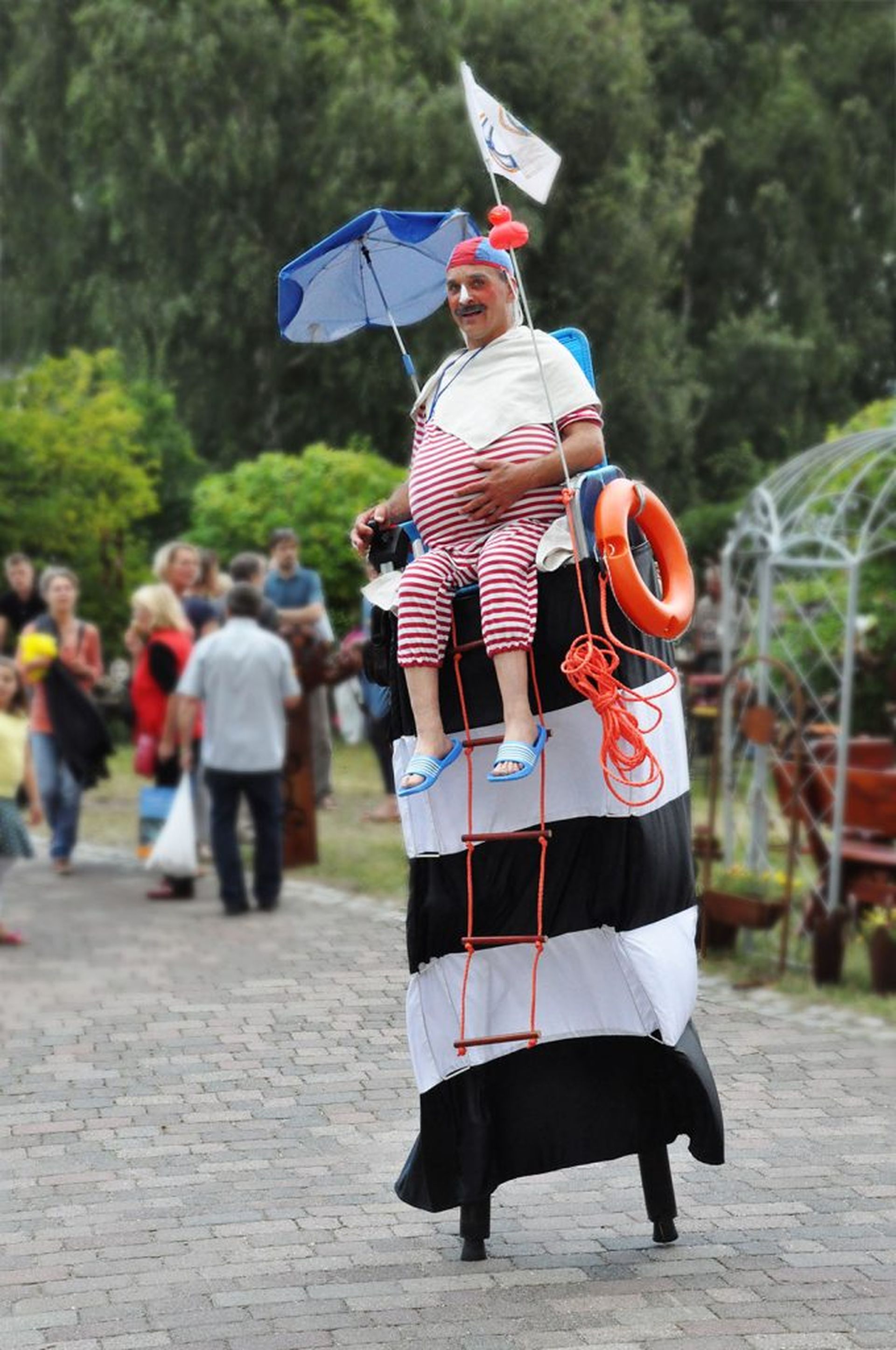 Jünni Bademeister geht auf Stelzen, die so verkleidet sind, dass es scheint als säße er auf einem Hochsitz, er trägt einen rot-weißen Badeanzug im Stil der 50er, hat einen Sonnenschirm und einen Rettungsring bei sich.
