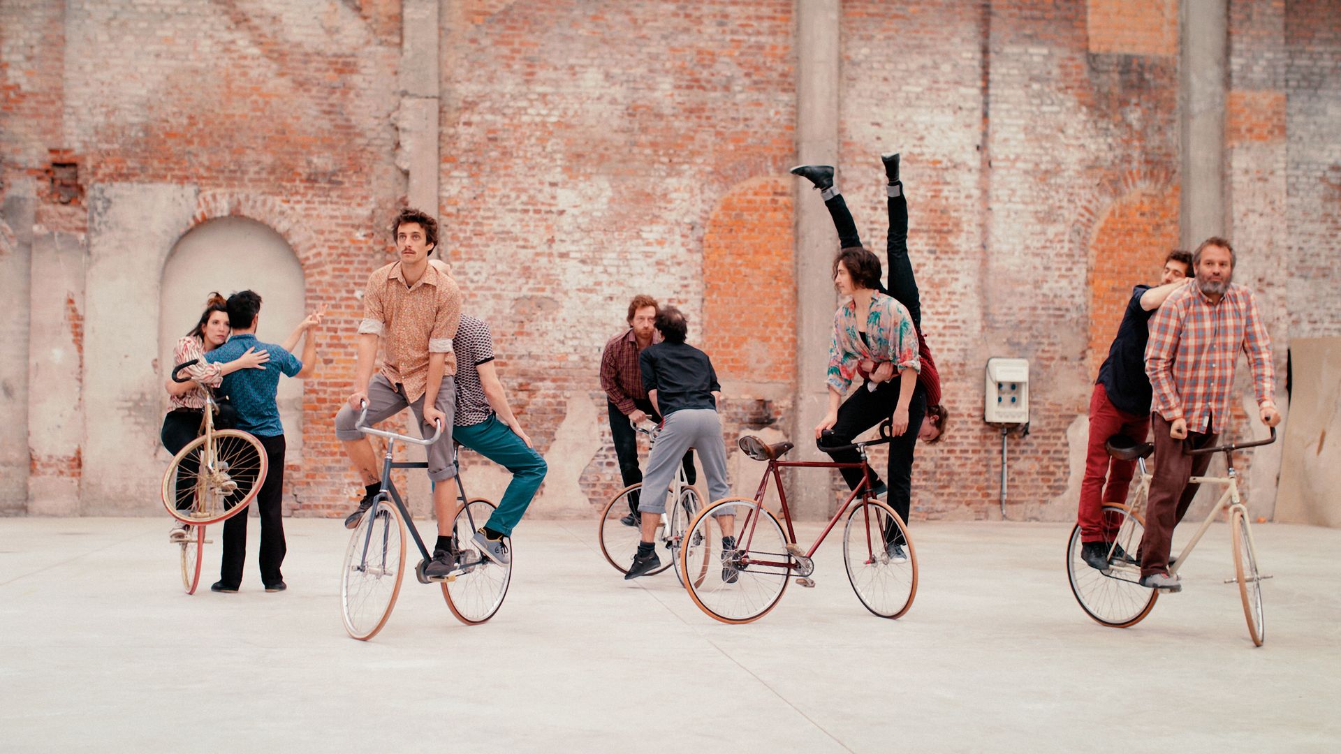 Das Foto zeigt zehn Personen, die jeweils zu zweit auf fünf Fahrrädern über einen Hof aus Schotter fahren. Im Hintergrund befindet sich ein Backsteingebäude. Manche fahren auf dem Rückrad, manche rückwärts, eine Person hält sich kopfüber am Partner fest