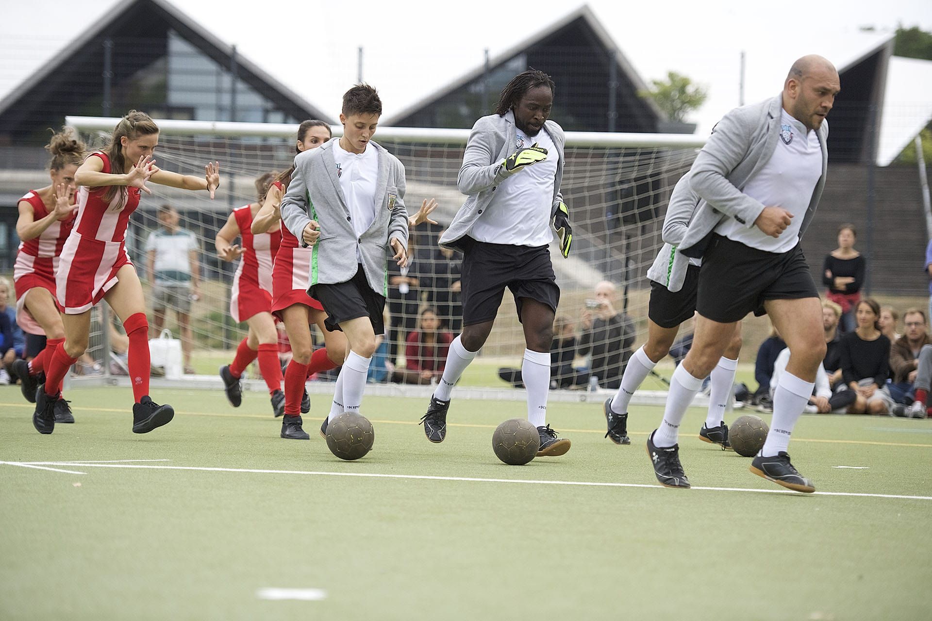 Im Hintergrund steht ein Tor, der Boden ist grüner Teppich. Durch das Bild laufen Fußballspieler*innen. Die eine Mannschaft trägt weiß-rote Trikot-Kleider, die andere schwarz-weiße Trikots und ein graues Jackett. Die Mannschaften scheinen nach Geschlechte