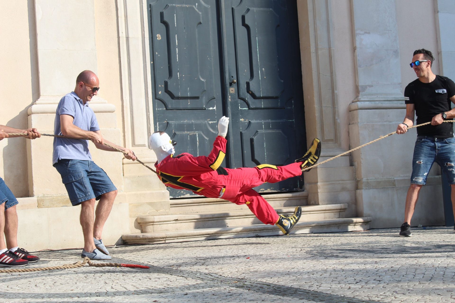 Der Clown Murmuyo, in rotem Overall mit schwarz-gelben Applikationen, schwebt in der Vertikalen, von einem Seil getragen, das Passant*innen halten.