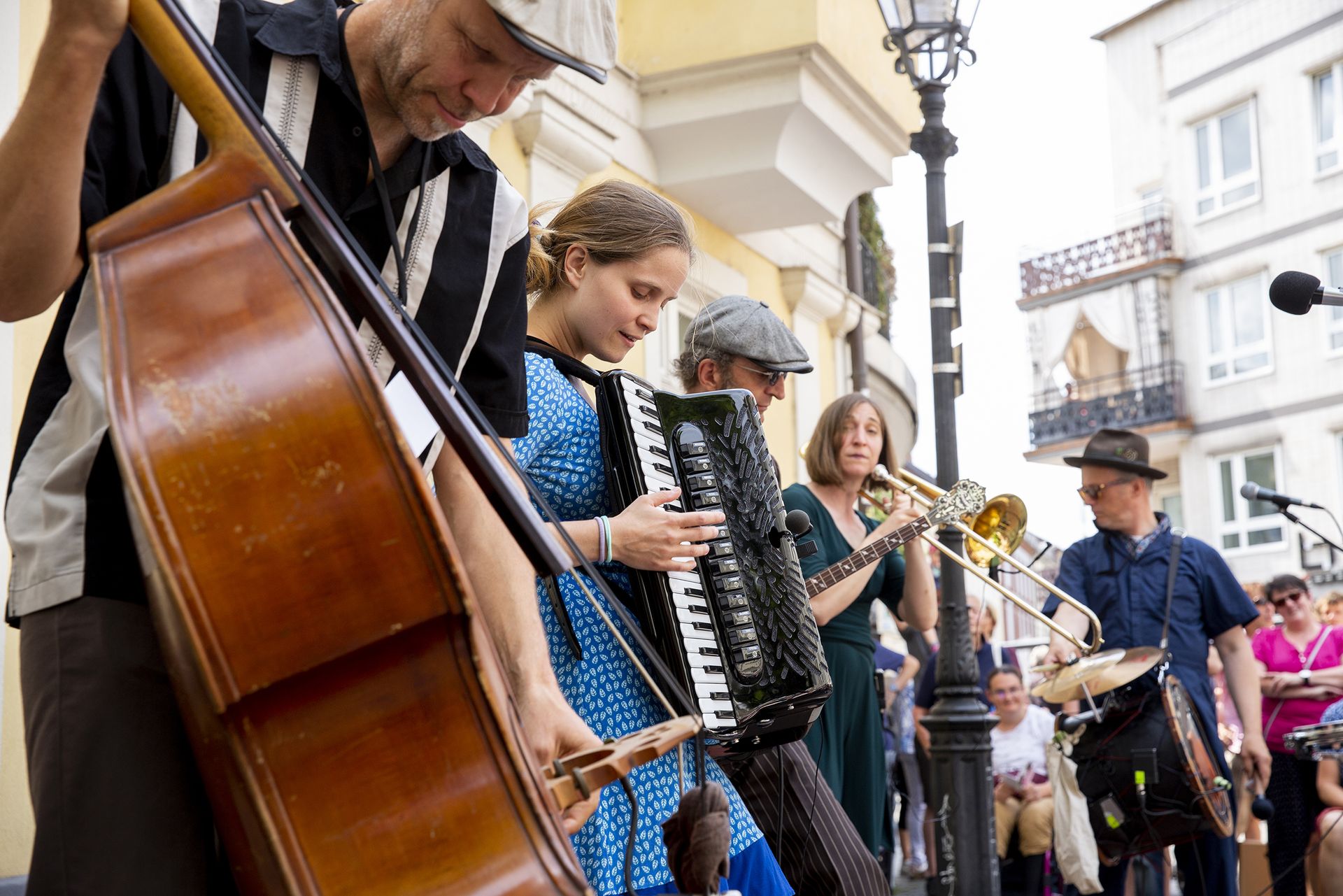 Die Person links steht am nächsten an der Linse, während die nächsten vier sich langsam von links nach rechts verlaufen von der Linse entfernen. Die Musiker*innen spielen Kontrabass, Akkordeon, Banjo, Posaune und Drum Kit.