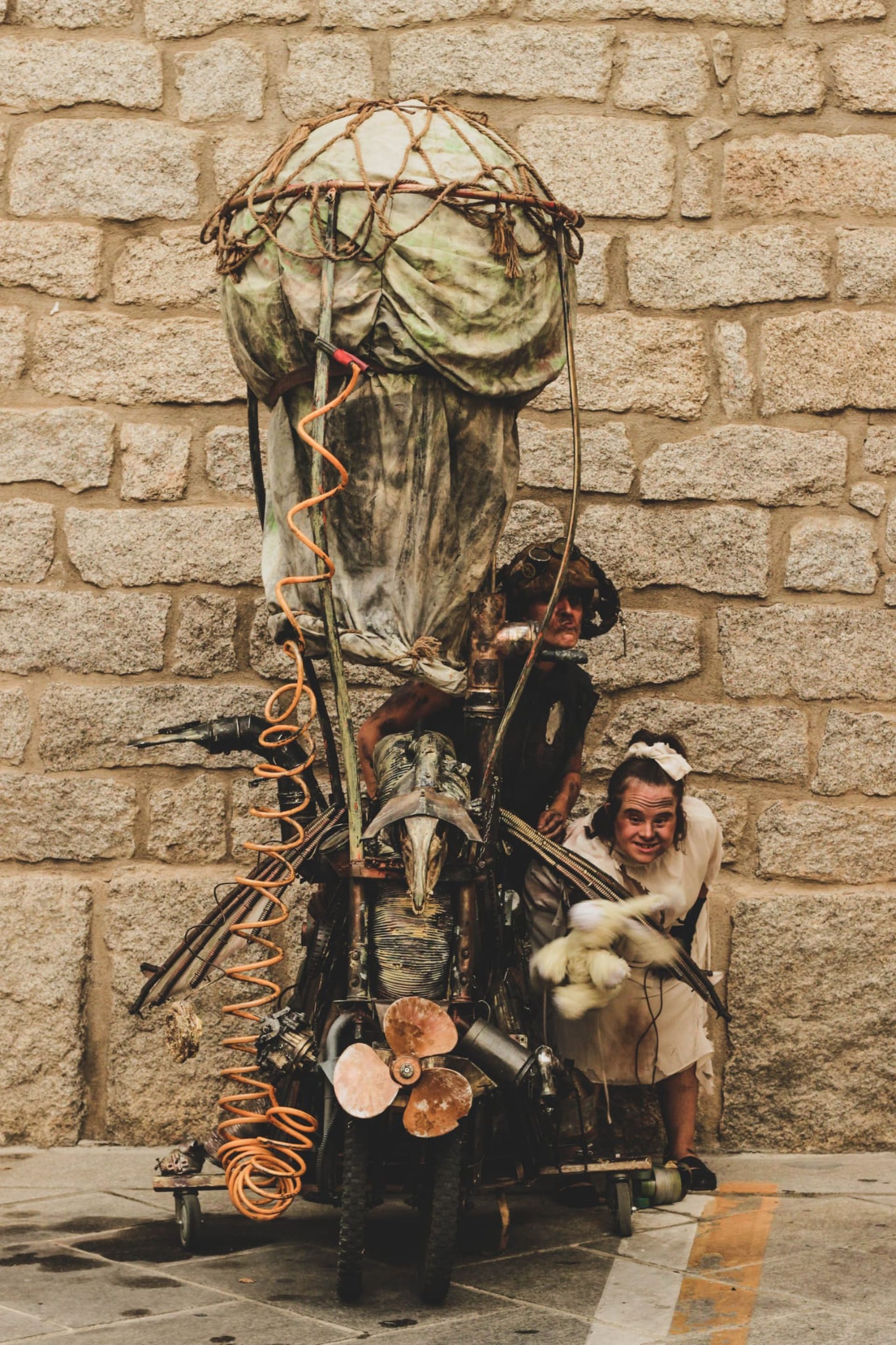 Zwei Personen und ein Fahrrad, das so hergerichtet ist, als sei es ein Motorrad. Die Menschen stehen rechts vom Gefährt. Kostüm und Dekoration sind dem Steampunk entnommen, wie bei Mad Max oder dem Videospiel BioShock. Rostige Technik, apokalyptisch.