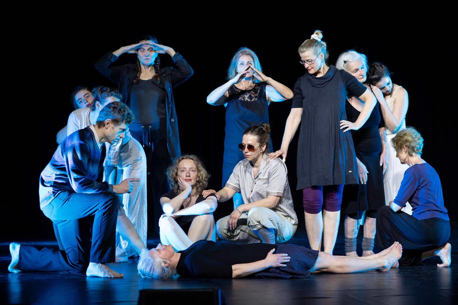 Gruppe von zwölf Menschen, in einer Position verharrend. Die Beleuchtung ist bläulich eingefärbt. Die Personen sind auf räumliche Ebenen verteilt: eine liegt, drei knien, zwei stehen gebeugt, fünf aufrecht.