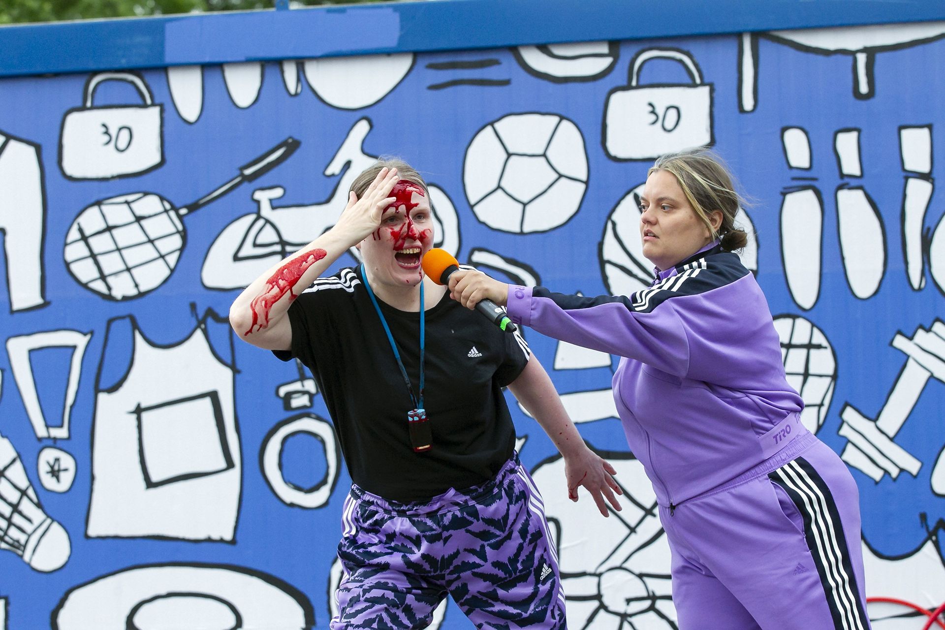 Zwei Performer*innen vor einer blau-weiß besprühten Wand. Sie tragen lila Sportkleidung. Die Person links ist überströmt mit Kunstblut, die Person rechts hält ihr ein Mikrofon vor den Mund.