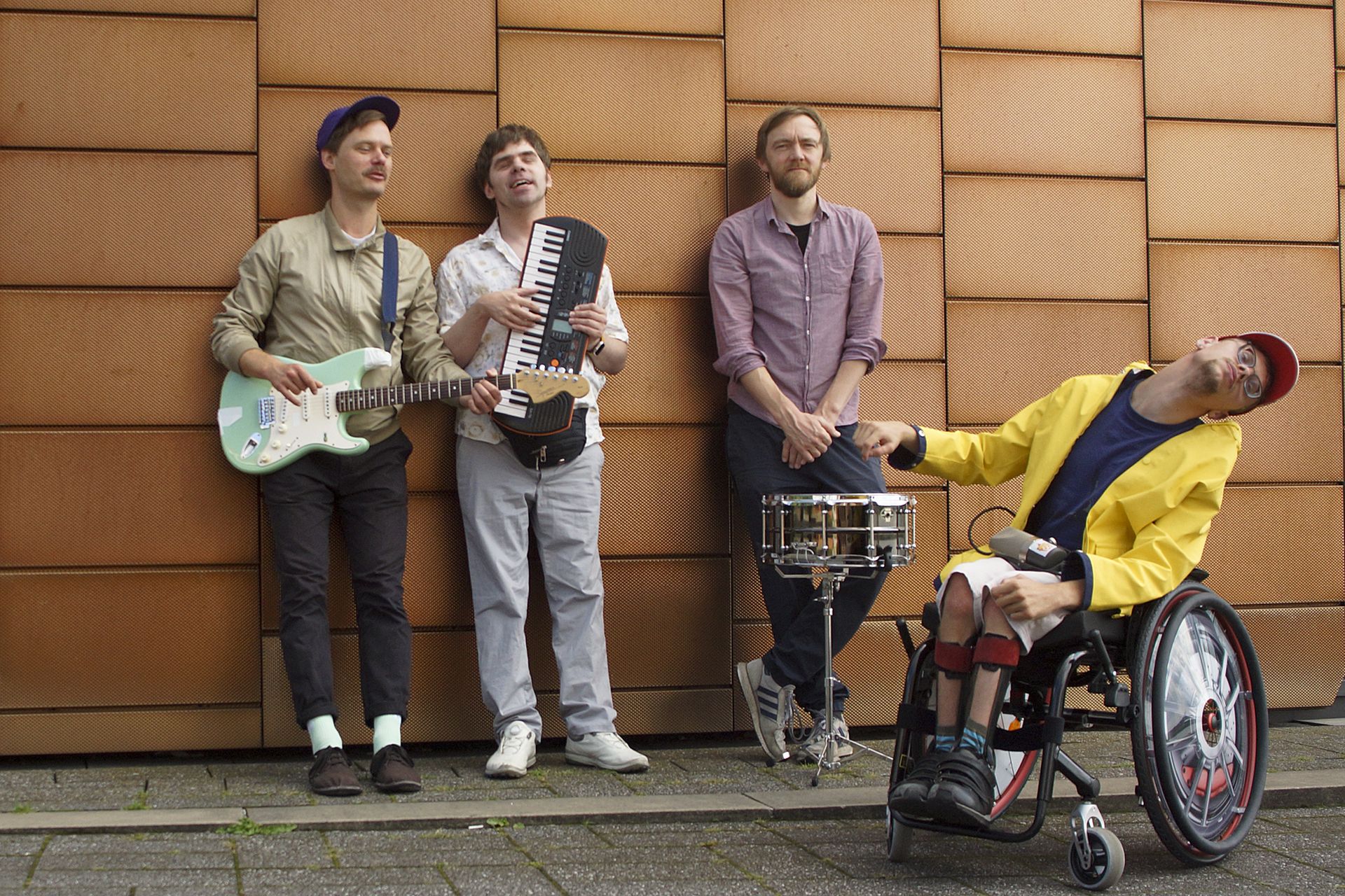 Vier Musiker lehnen lässig vor einer kupferfarbenen Wand: v.l.n.r. Person mit weiß-grüner elektrischer Gitarre, Person mit Keyboard, Person mit Drumkit vor sich, Person mit Rollstuhl und Mikrofon im Schoß.