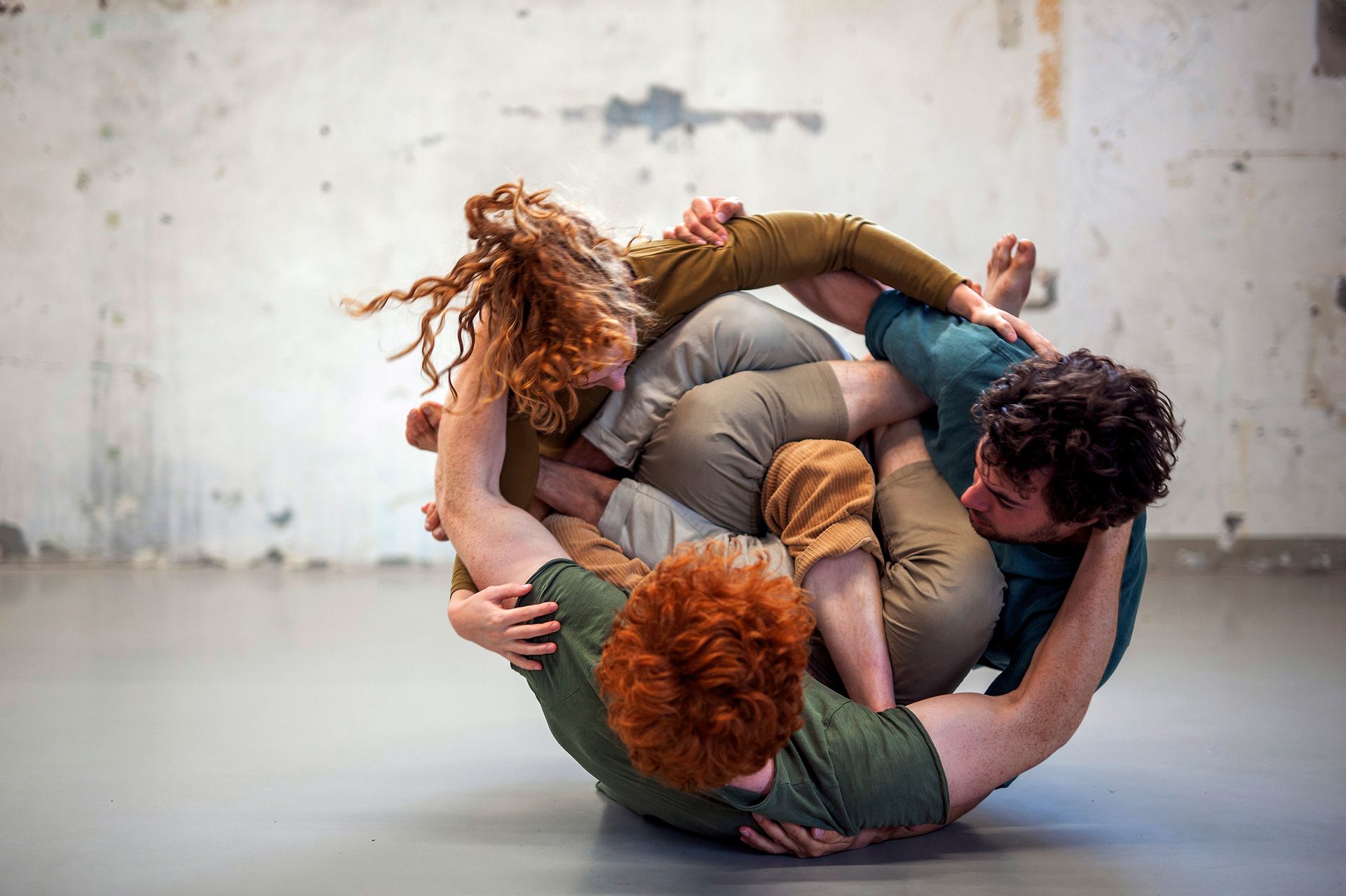 Drei Artist*innen haben die Beine so verknotet und die Arme so fest umeinander geschlungen, dass sie wie ein Ball aus Menschen scheinen und sich rollend durch das Bild bewegen.
