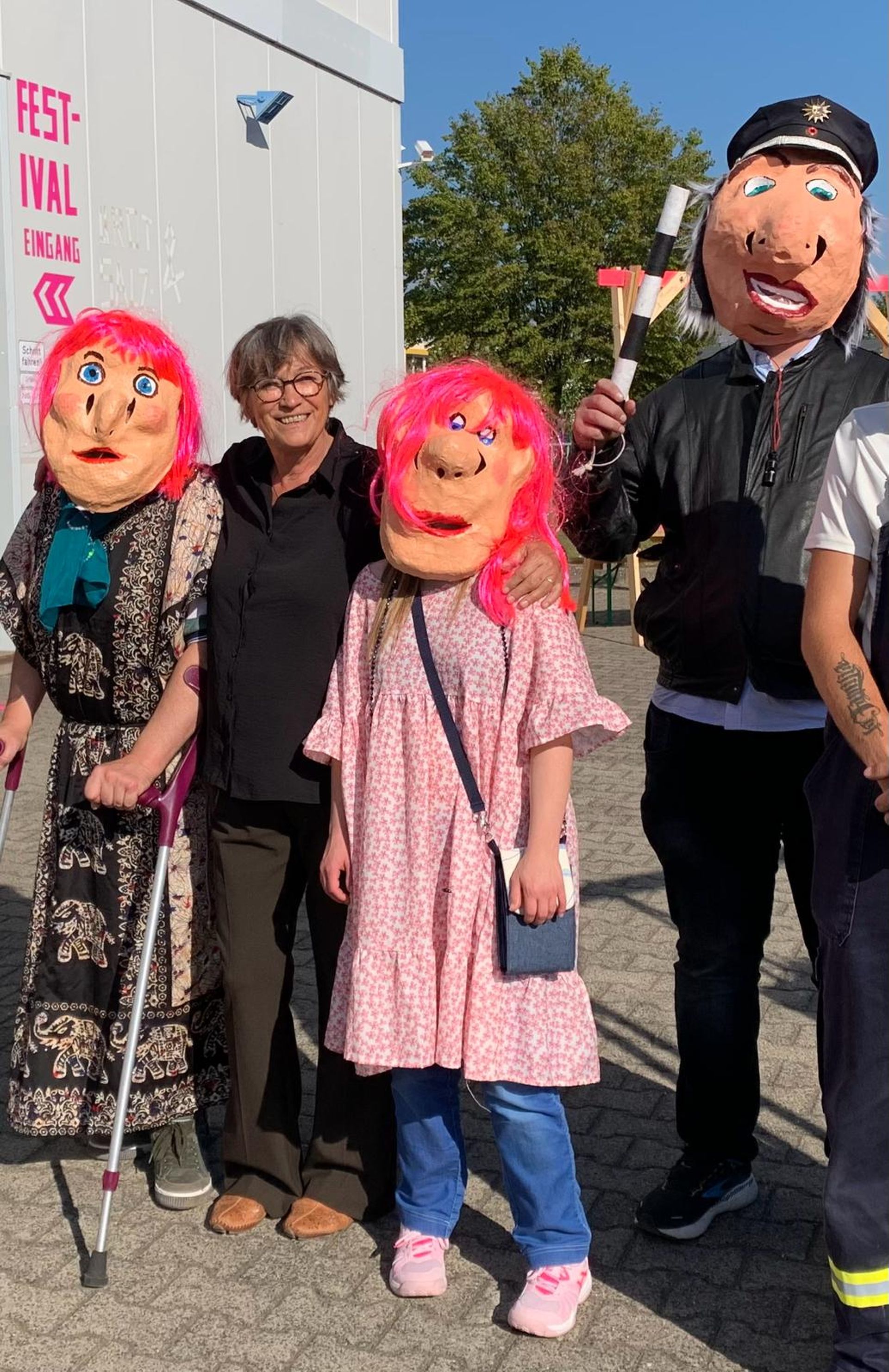 Vier Personen schauen in die Kamera, drei von ihnen tragen selbstgemachte Maskenköpfe. Zwei der Masken haben knallpinke Haare, der dritte trägt den Hut einer Polizeiuniform.