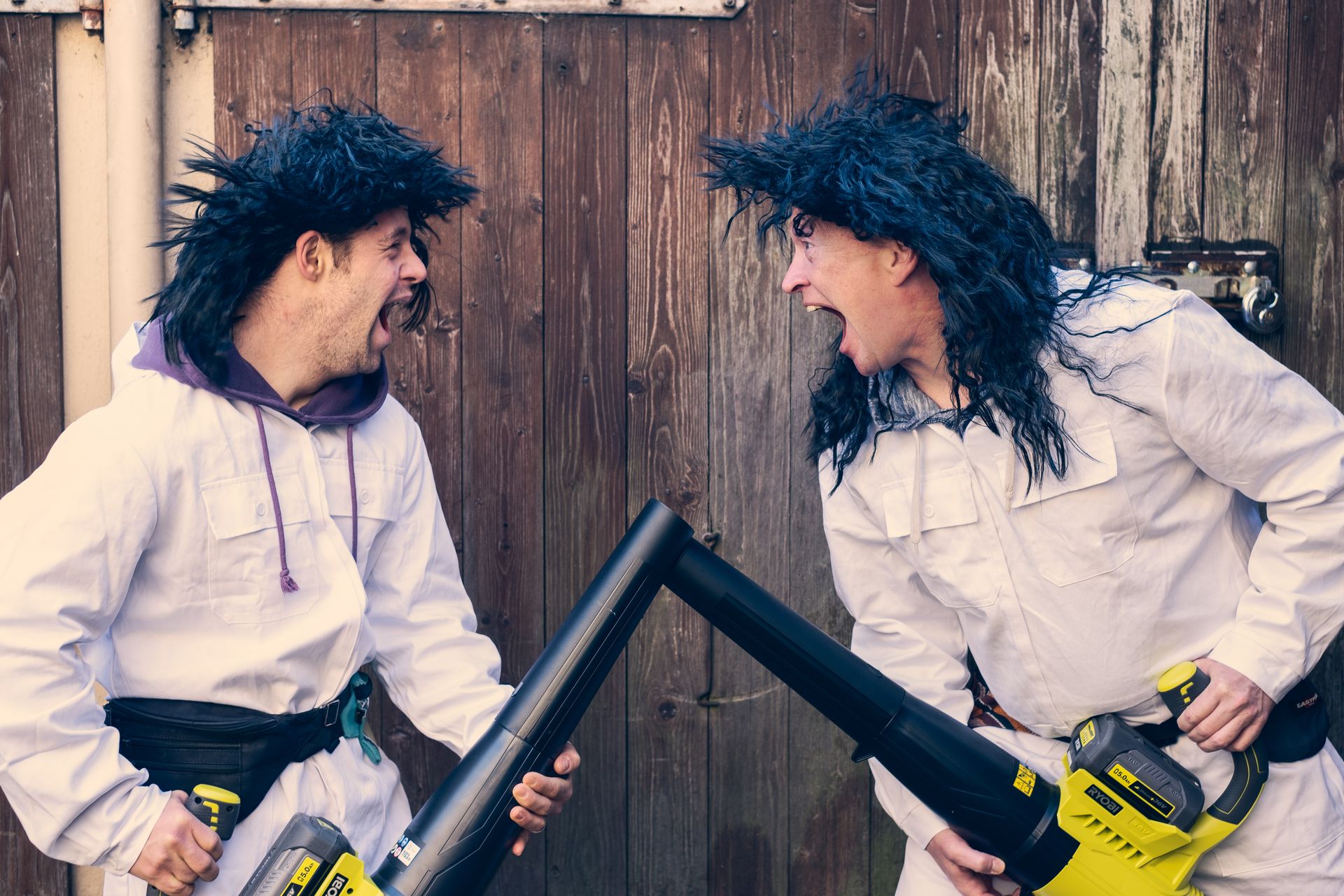 Zwei Personen stehen sich gegenüber und schreien sich an. Sie tragen weiße Overalls, schwarze zottelige Perücken und haben Laubgebläse in der Hand, die sie aufeinander gerichtet haben.